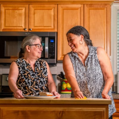 Senior woman and her caregiver smiling at each other while serving food on a plate.