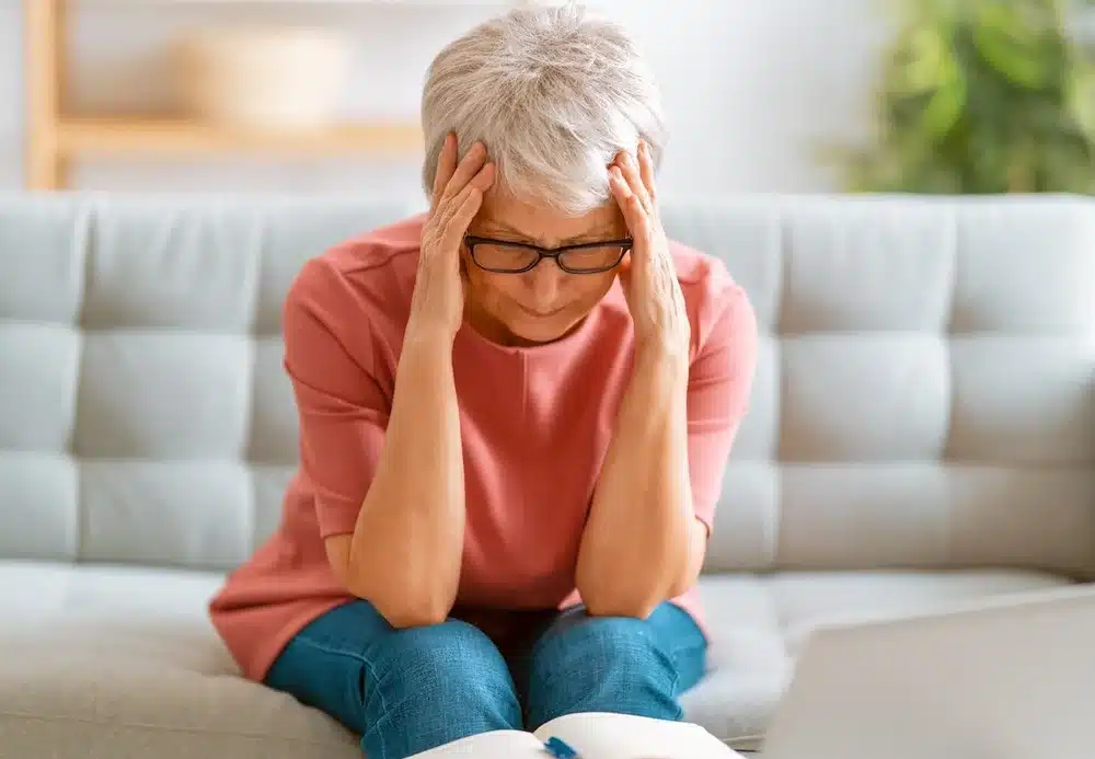 Elderly woman with short gray hair and glasses sits on a sofa, holding her head in frustration, a book open in front of her, expressing stress.