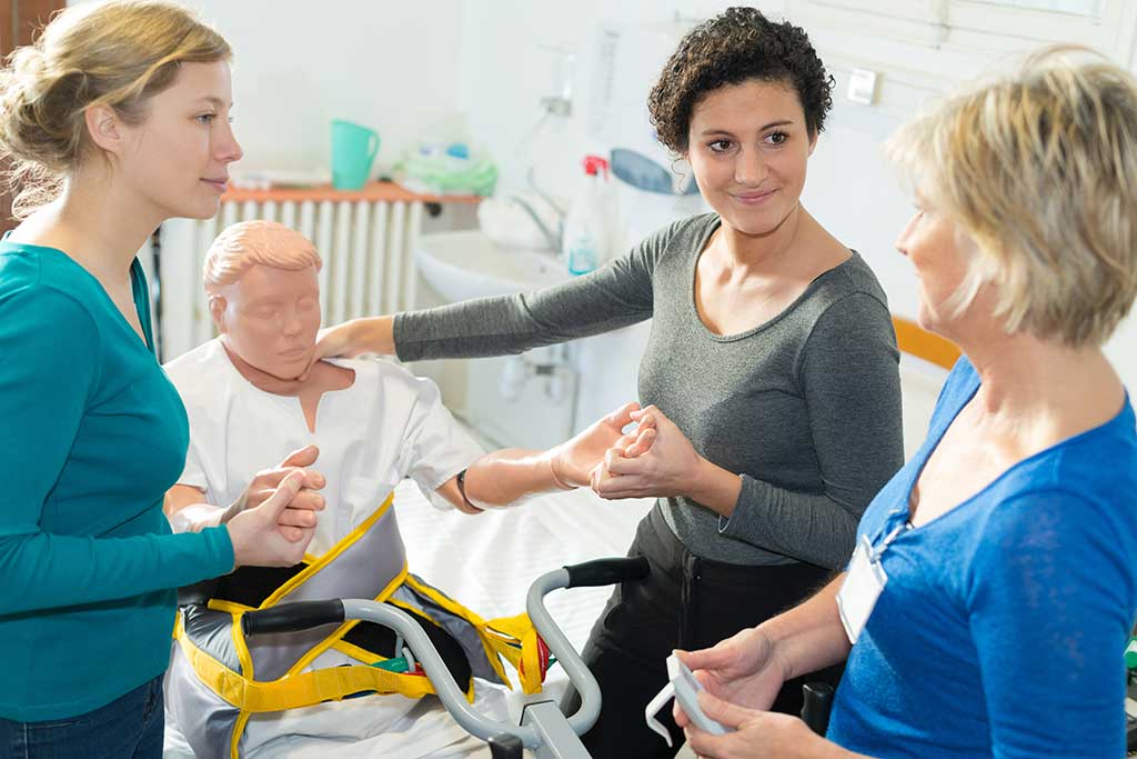 Three women practice patient lifting using a medical mannequin in a healthcare setting. They appear focused and collaborative, conveying teamwork.