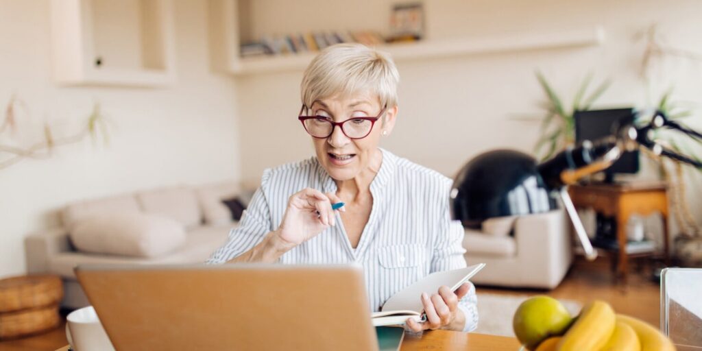 Elderly woman with glasses holding a notebook, looking surprised at a laptop in a cozy living room. The scene conveys curiosity and excitement.