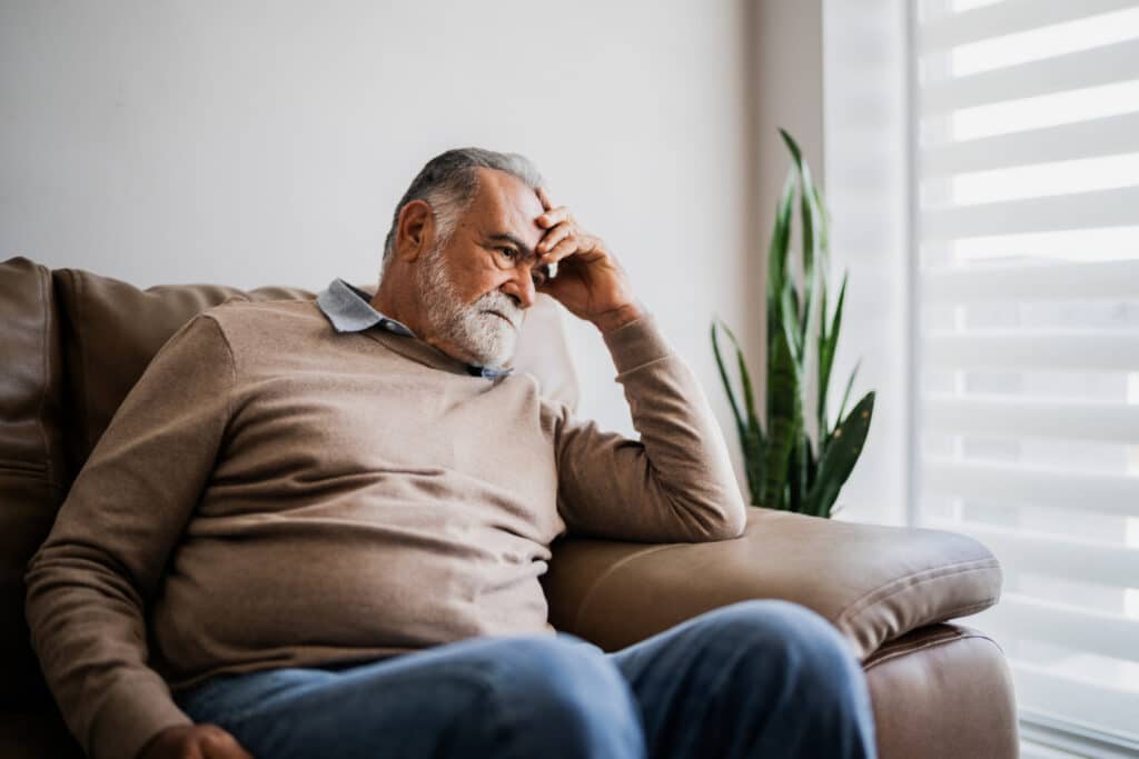 An elderly man with a beard sits pensively on a brown leather couch, hand on head. Soft light filters through blinds, with a plant nearby, conveying contemplation.