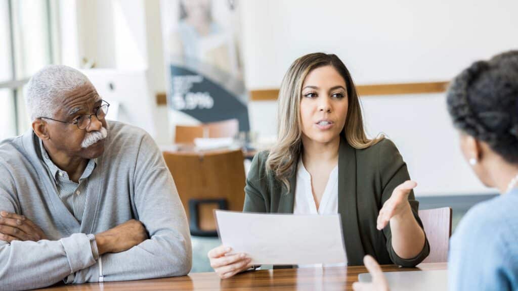 A woman in a green blazer holds a document and gestures as she speaks with an older man and a colleague in a bright, collaborative office meeting.