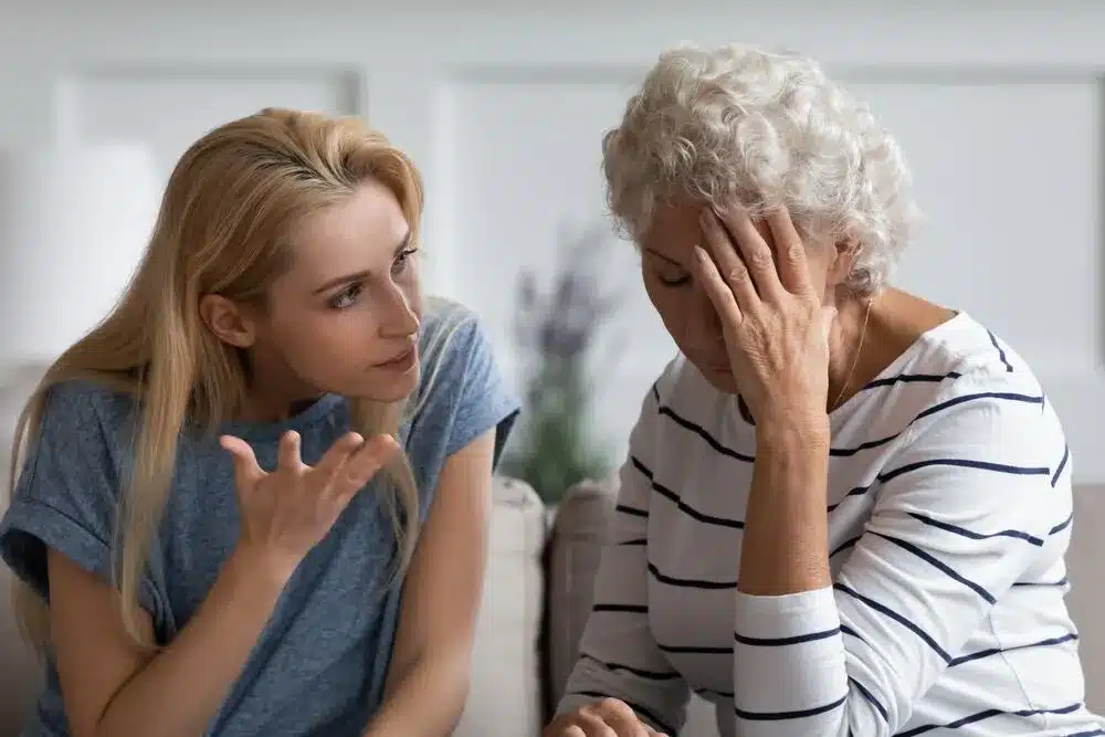 Concerned elderly woman sitting with her hand on her forehead while a younger woman berates her beside her in a home setting. Emotional conversation scene representing senior mental health, stress, family support, caregiving, anxiety, depression, empathy, counseling, and intergenerational communication. Soft indoor lighting with a calm, intimate atmosphere.