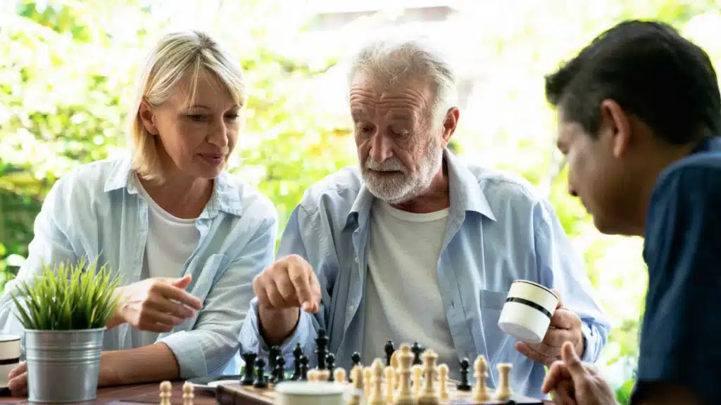 Older man playing chess with two companions outdoors, illustrating cognitive engagement, social interaction, and brain-stimulating activities for healthy aging.