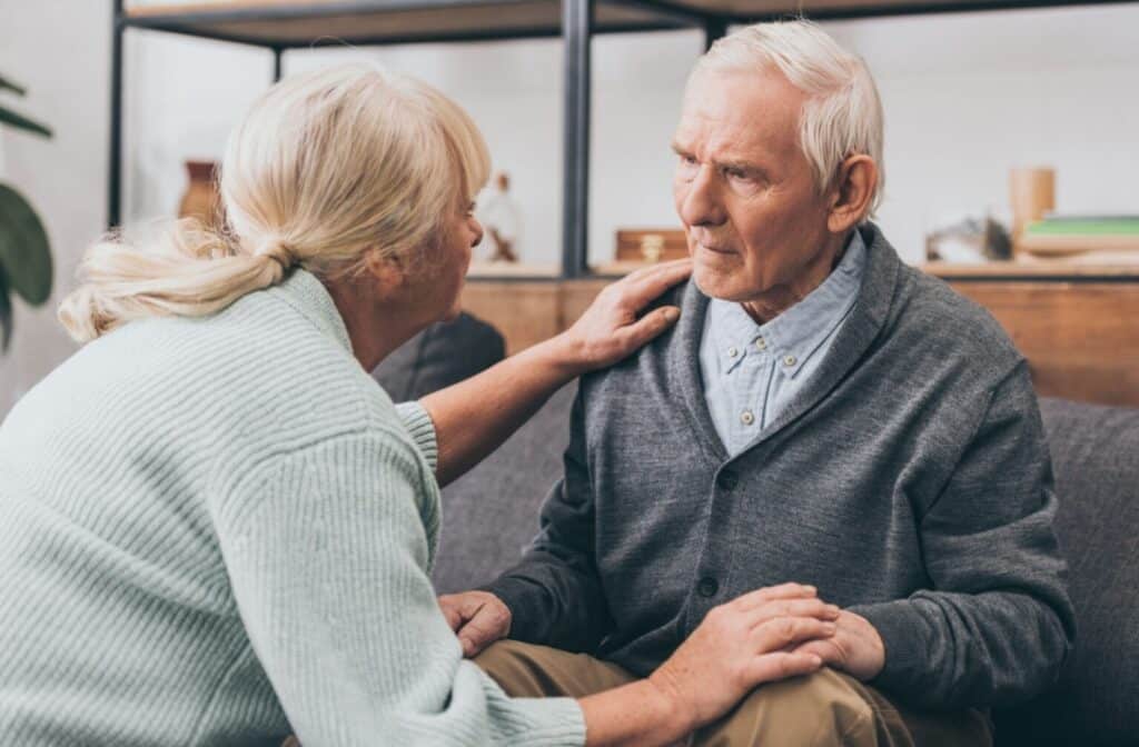 An elderly woman, wearing a light blue sweater, gently consoles an elderly man in a gray cardigan, conveying empathy and concern in a domestic setting.