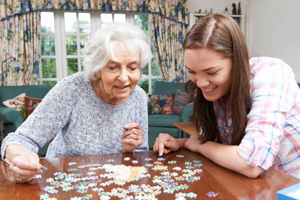 An older woman and younger woman smile together while assembling a jigsaw puzzle at a table in a bright, cozy living room.