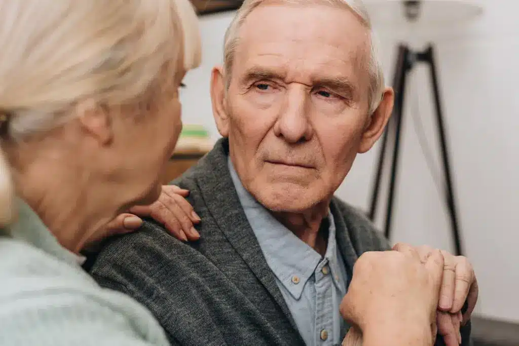 Elderly man in a gray cardigan gazes thoughtfully at a woman beside him, who rests a reassuring hand on his shoulder in a caring scene.