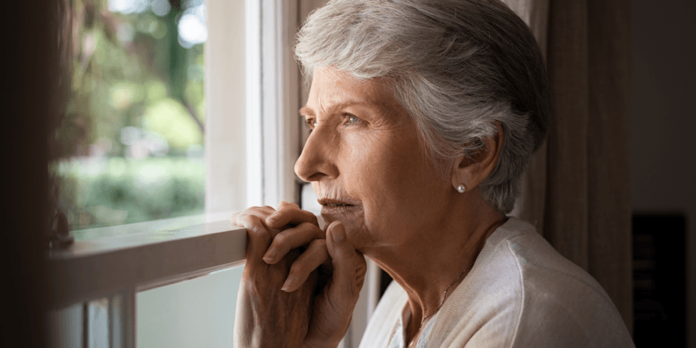 Older woman with short gray hair gazes thoughtfully through a sunlit window, resting her hands on the sill; calm, reflective mood.