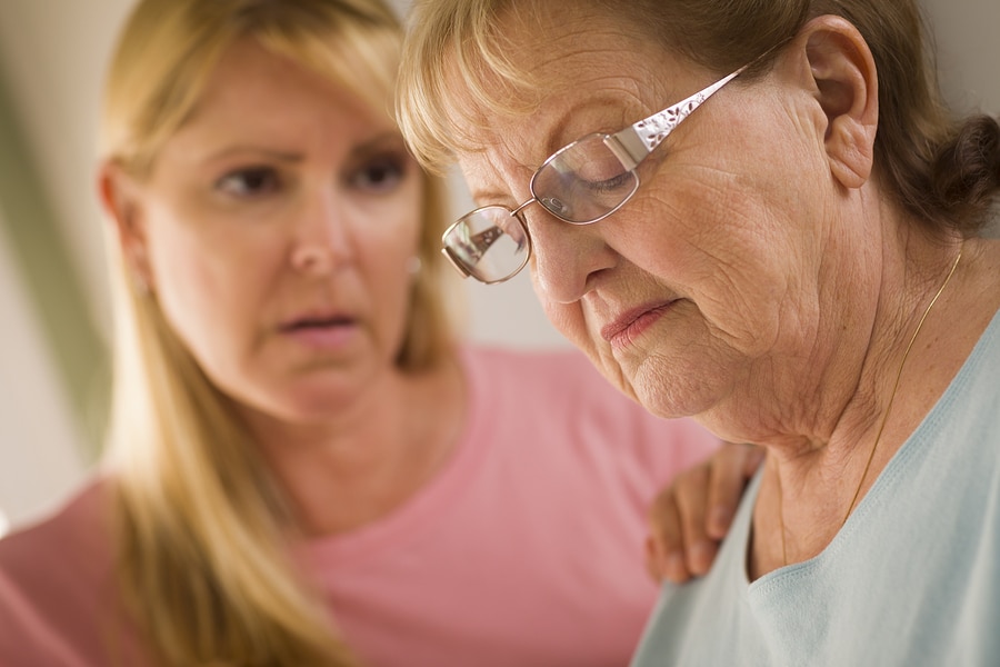 An older woman in glasses looks down with a worried expression as a younger woman in pink rests a hand on her shoulder, offering support.
